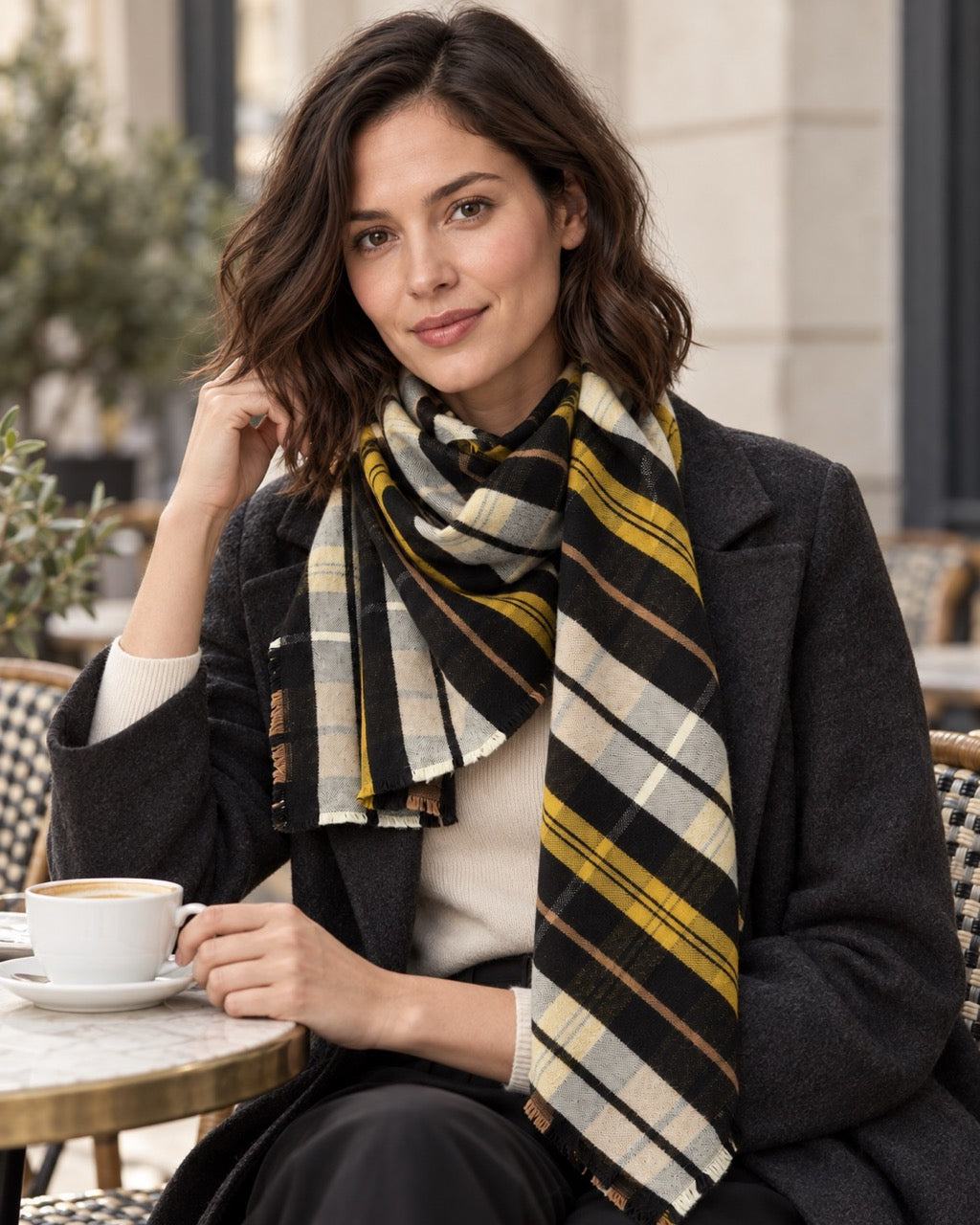 Model seated at a café terrace wearing an s.Oliver multicolour checked scarf with short frayed edges, a charcoal coat and cream top.